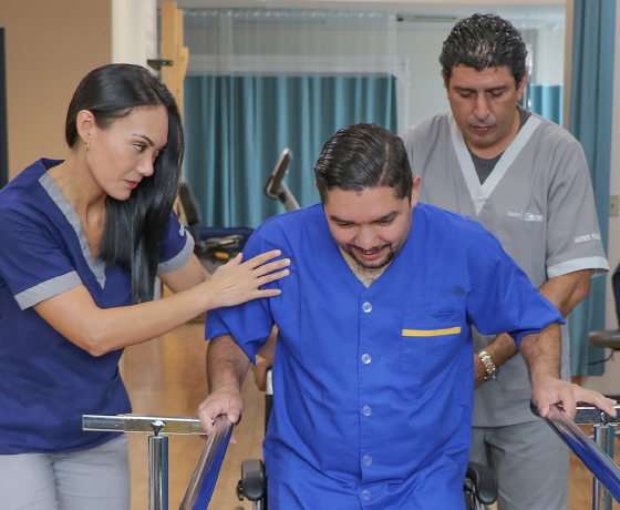 Terapeuta y asistente ayudándole a paciente a caminar sobre gradas de madera. La terapeuta la guía por el frente y la asistente la acompaña por la espalda.